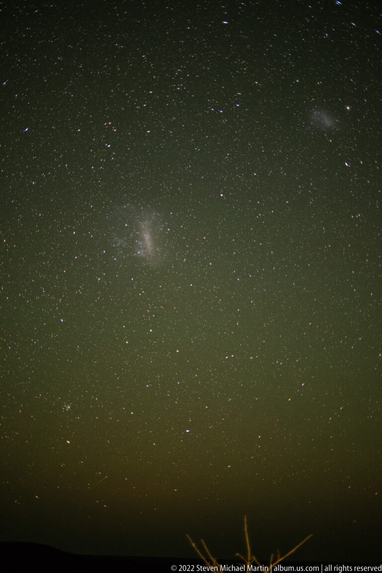 Dense southern star field with two diffuse luminous cloudlike patches above a narrow dark horizon.
