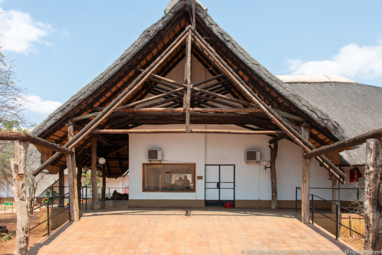 Front of a low white Shingwedzi camp building beneath a steep thatched gable and exposed timber trusses.