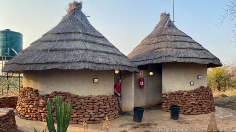 Two round Punda Maria camp buildings with conical thatched roofs, earth-toned walls, and reddish stone bases.