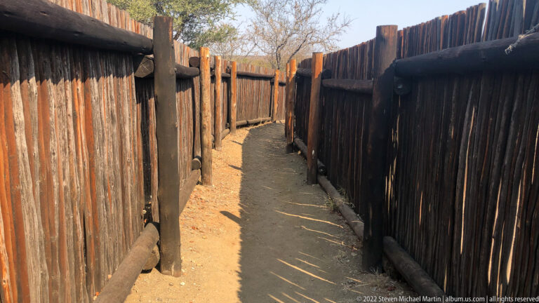 Narrow sandy walkway enclosed on both sides by tall vertical timber-pole fencing.