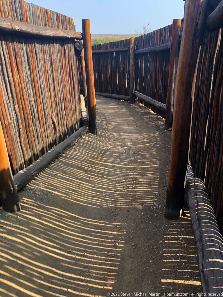Curving screened wooden walkway with strong striped shadows from vertical pole walls.