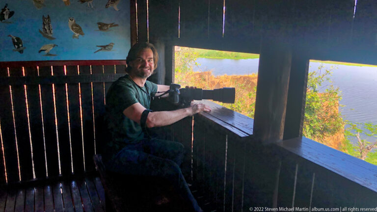 Seated man using binoculars through a narrow opening inside the dark wooden wildlife hide.