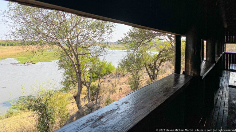 Waterhole and shoreline trees seen through the dark wooden opening of Matambeni Hide.