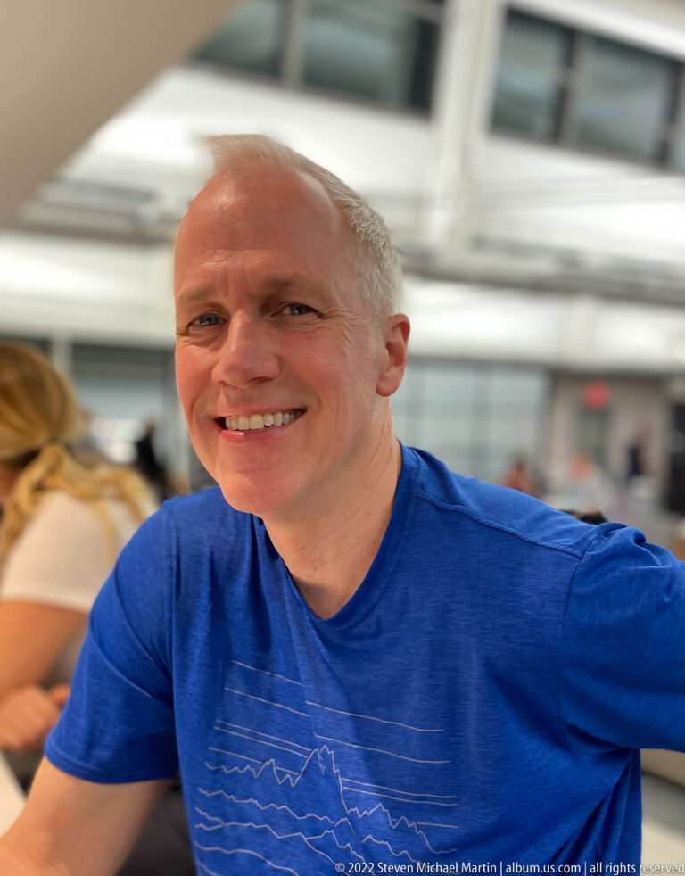 Smiling traveler in a blue shirt seated at a table in a bright terminal interior.