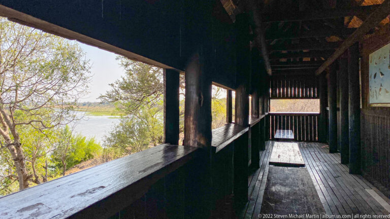 Dark wooden bench and viewing opening inside Matambeni Hide overlooking the bright waterhole.
