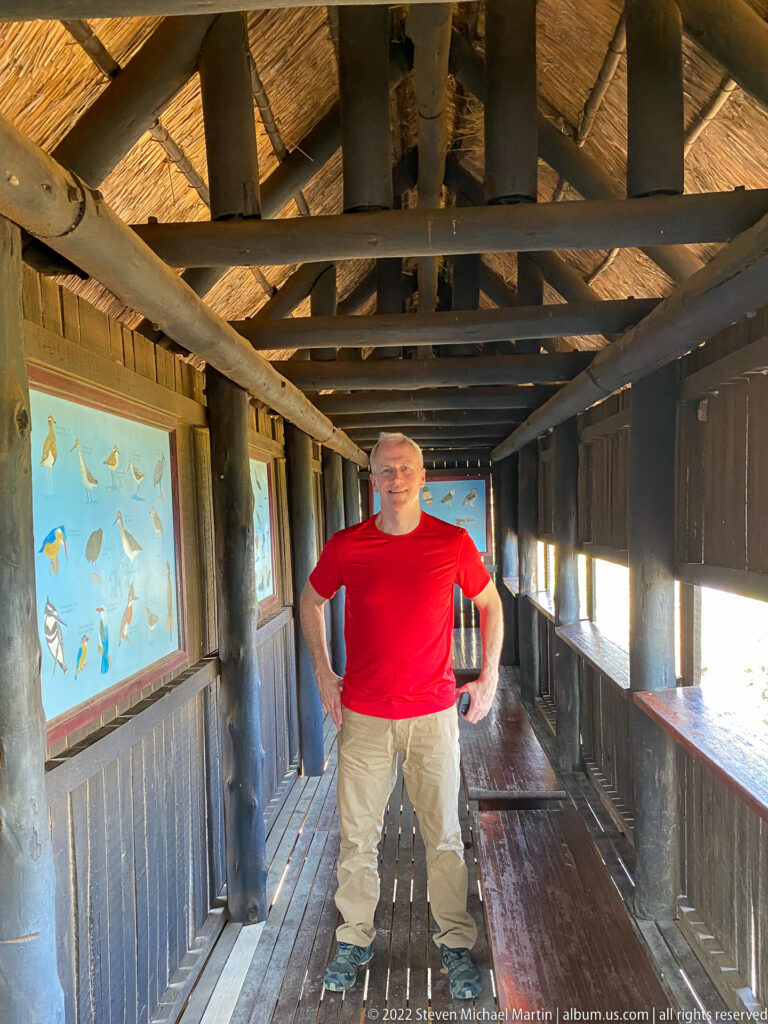 Man standing inside a long wooden wildlife hide with benches, viewing slots, and bird-identification panels.