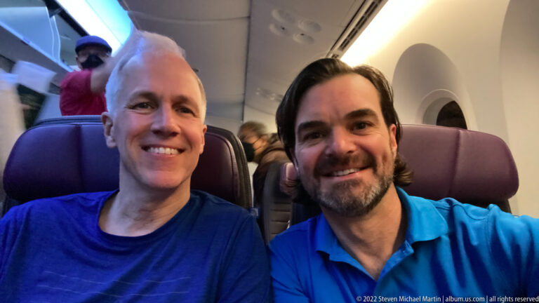 Two smiling travelers seated together in high-backed seats inside an aircraft cabin.