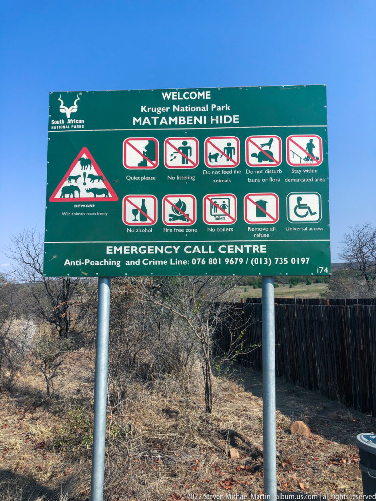 Green Matambeni Hide sign listing wildlife-viewing rules and emergency contact information beside dry woodland.