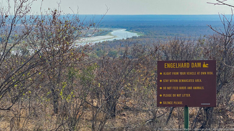 Engelhard Dam rules sign in the foreground with the distant dam and dry woodland visible beyond.