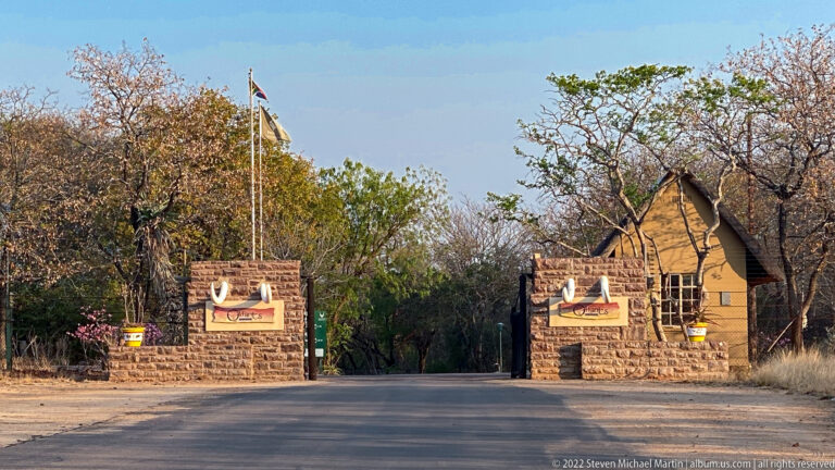 Road entrance to Olifants Rest Camp with stone gate walls, tusk signs, and a lodge building beyond.