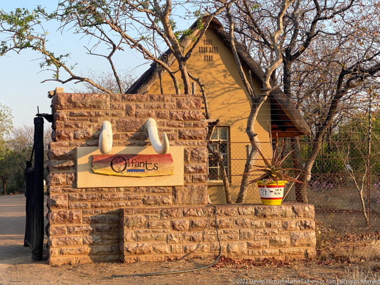 Stone entrance monument labeled Olifants in front of a steep-roofed lodge-style building and dry woodland.