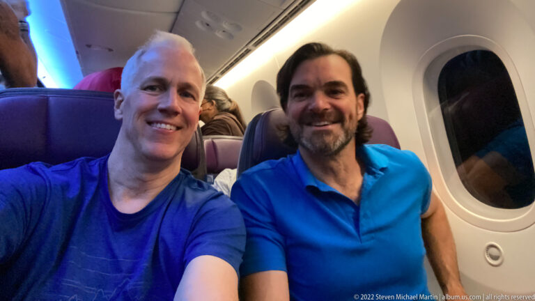 Two smiling travelers seated beside an aircraft window with other passengers visible behind them.