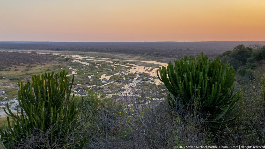 Elevated dusk view over a braided river with rocky channels, sandbars, bushveld, and tall succulents in the foreground.