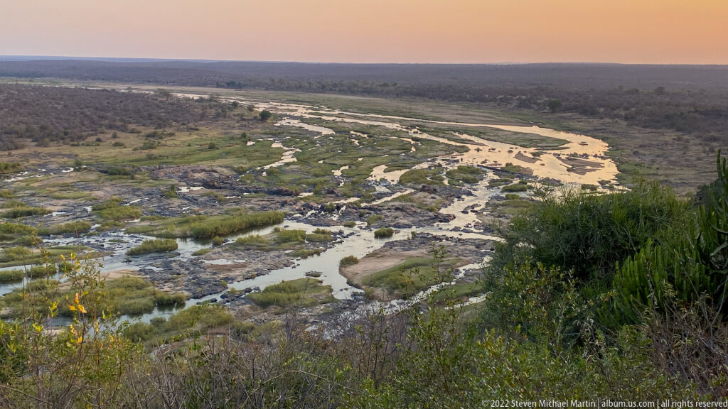 Wide elevated view of shallow braided river channels crossing rock, sand, and green vegetation in dry bushveld.