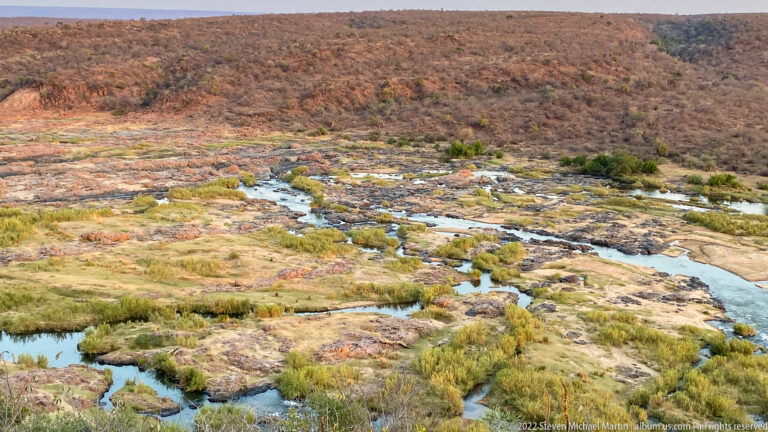 Elevated view of shallow braided water channels crossing exposed rock and vegetation below dry hills.