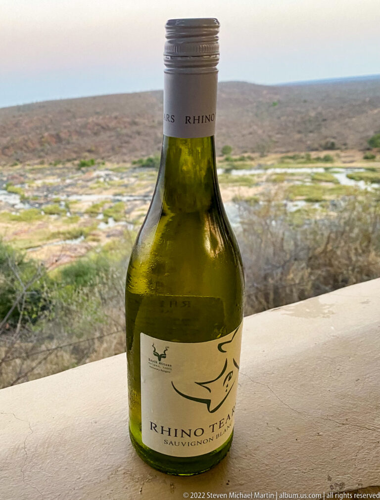 Green bottle labeled Rhino Tears standing on a ledge with a broad riverine landscape behind it.