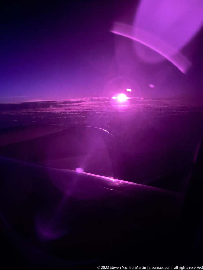 Dark nighttime view through an aircraft window showing a wing and engine beneath purple-magenta reflections.