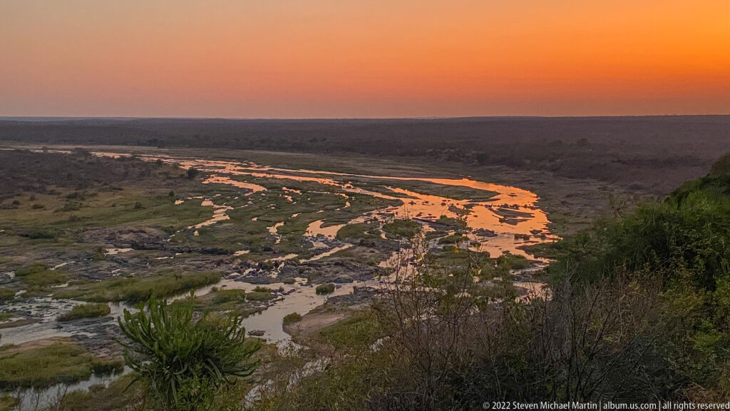 Braided river channels glowing orange beneath a vivid sunset sky, viewed from a high bushveld overlook.