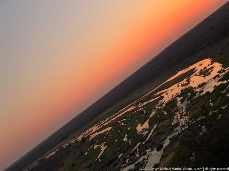 Elevated sunset view across reflective wetland channels and pools beneath an orange sky.