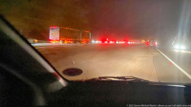 Night highway seen through a windshield with trucks, red taillights, and bright oncoming headlights.