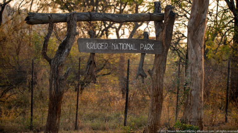 Rustic wooden sign reading Kruger National Park beside a wire fence in dry woodland.