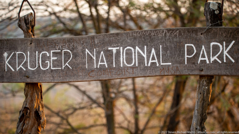 Weathered wooden sign reading Kruger National Park mounted between rough posts in woodland.