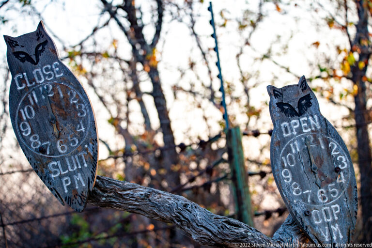 Weathered metal OPEN and CLOSE signs with clock numerals hang from a branch at a wooded camp entrance.