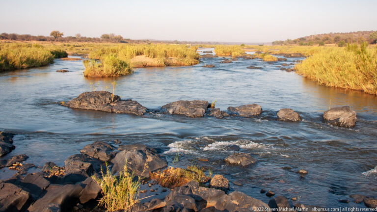 Shallow river flows around dark rocks and grassy islands, with reeds lining the banks under warm light.