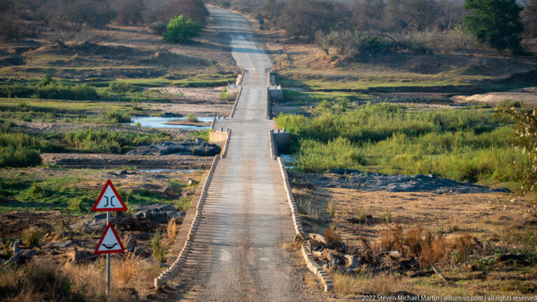 Long narrow concrete bridge crosses a broad shallow riverbed, with triangular wildlife-warning signs in the foreground.