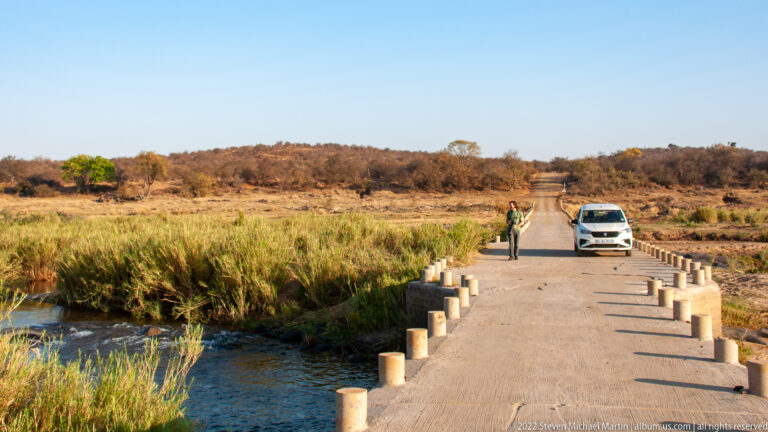 Narrow concrete bridge crosses a shallow river, with a parked safari vehicle and a person standing on the roadway.