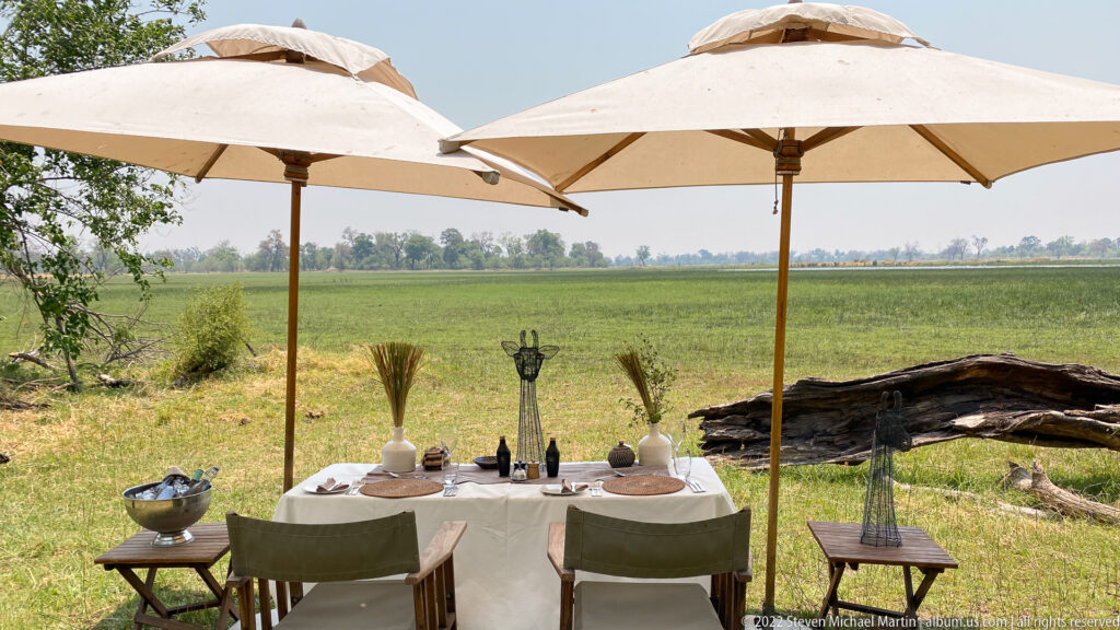Outdoor dining table set beneath canvas parasols overlooking a broad green floodplain.