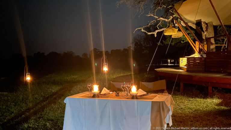 Candlelit table set for dinner on grass beside a raised safari camp deck at night.