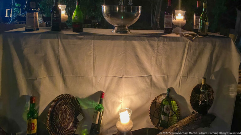 Nighttime drinks station with bottles, woven covers, candle lantern, and a metal bowl on a draped table.