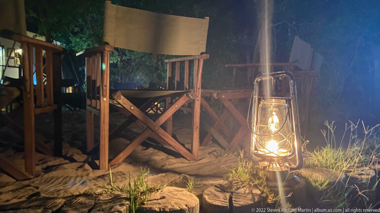 Glowing kerosene-style lantern beside wooden director’s chairs on sandy ground at night.