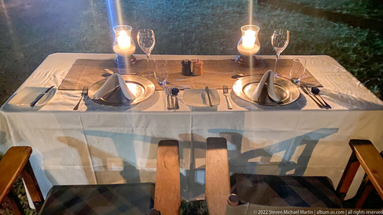 Table for two set with plates, folded napkins, wine glasses, cutlery, and candle lanterns outdoors at night.