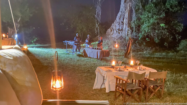 Several candlelit dining tables arranged on grass beneath large trees at a safari camp at night.
