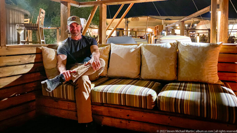Adult guest seated on a striped cushioned wooden bench inside an open-sided safari lodge.