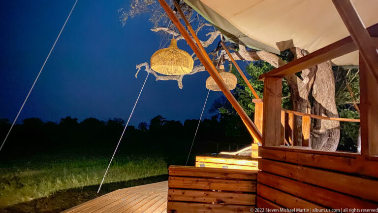 Raised wooden safari-camp deck with canvas roof and hanging woven lamps at dusk.