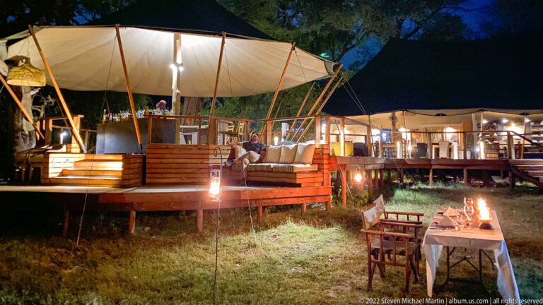 Illuminated open-sided safari lodge with timber decks, canvas roofs, and a candlelit dining table at night.