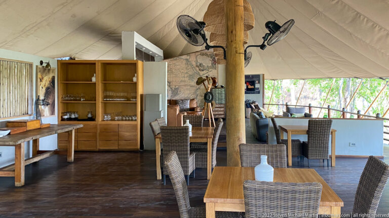 Open safari lodge dining area with wooden tables, woven chairs, shelving, bar counter, and canvas roof.