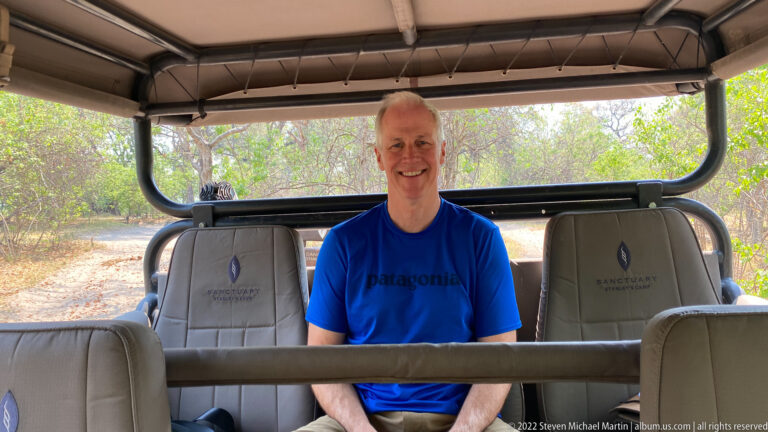 Smiling adult seated in the rear of an open safari vehicle with dry woodland behind.