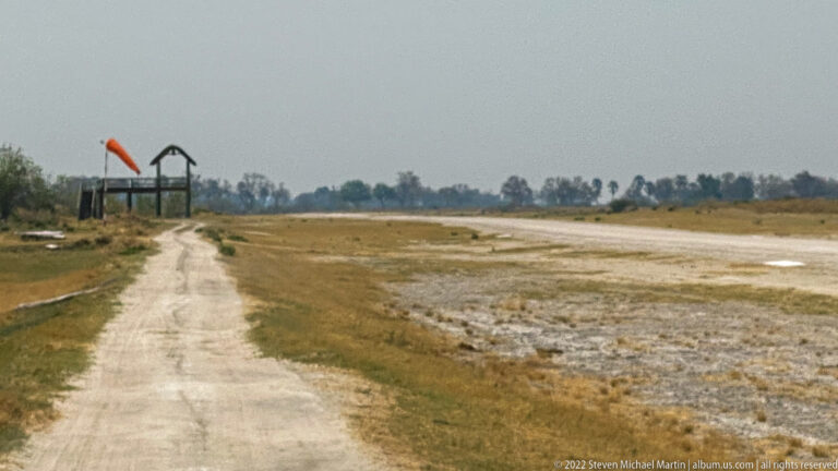 Unpaved bush airstrip stretching toward an orange windsock and simple raised structure.