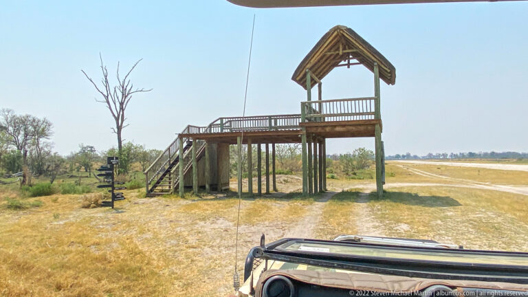 Raised wooden platform with stairs and thatched roof beside an unpaved safari airstrip.