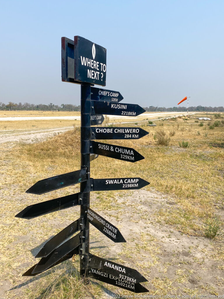 Black multi-arrow signpost listing safari camps and distances beside a pale bush runway.