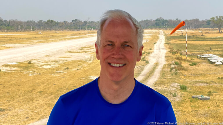 Smiling adult in a blue shirt at a bush airstrip with runway and orange windsock behind.