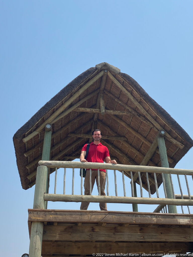 Adult guest standing behind the railing of a raised thatched airstrip platform, viewed from below.