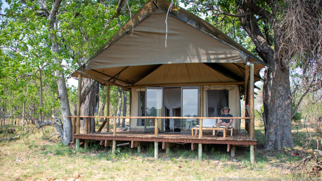 Raised canvas safari tent with wooden veranda beneath large trees in dry woodland.