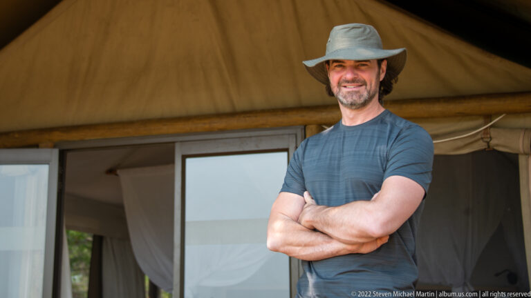 Smiling adult guest in a broad-brimmed hat standing outside a canvas safari tent.