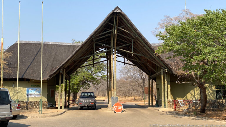 Sedudu Wildlife Gate entrance with tall timber gable, thatched roofs, vehicles beneath the canopy, and a red stop sign.