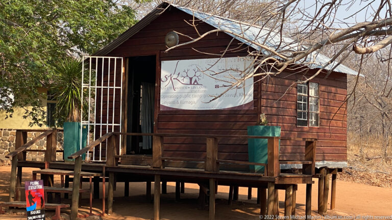 Small raised timber kiosk with porch, steps, barred doorway, and a large sign, standing beside dry woodland near the park entrance.